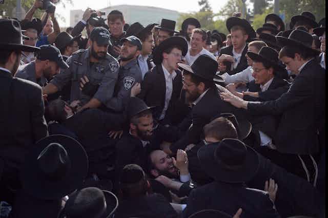 A crowd of several dozen people, most of them wearing white shirts, black jackets and black hats, scuffle with three men in grey uniforms.