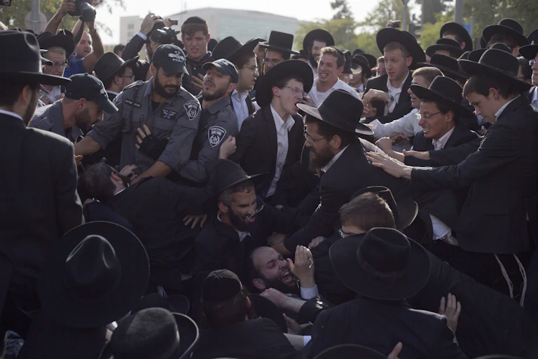 A crowd of several dozen people, most of them wearing white shirts, black jackets and black hats, scuffle with three men in grey uniforms.