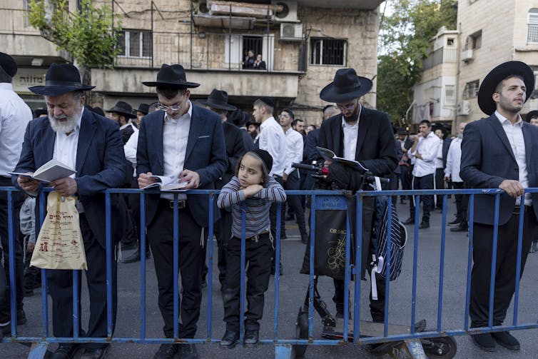 Four men in black hats and jackets, as well as a child, stand near a blue fence on a street, as they men look down at books in their hands.