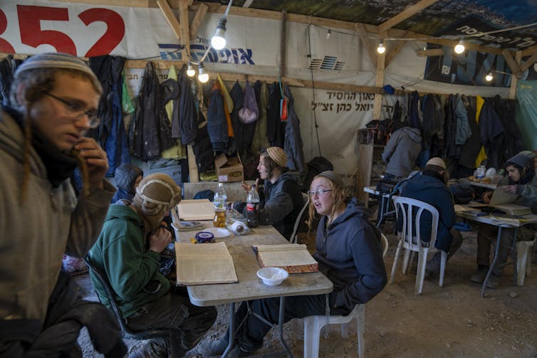 Young men sit at tables in a dimly lit temporary structure.