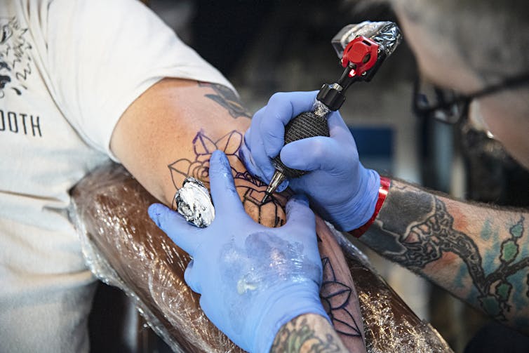 A tattoo artist wearing latex gloves holding a tattooing needle inks a geometric design on an arm.