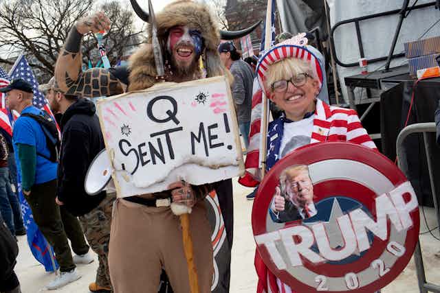 A man wearing red, white and blue face paint holds a sign that says 'Q sent me,' and poses with his arm raised next to a blonde woman who is wearing American flag clothing and holding a large, round banner that says 'Trump 2020.'