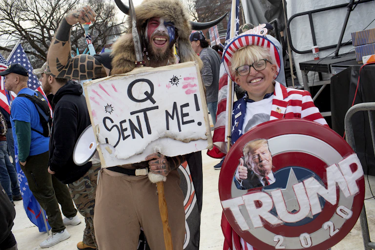 A man wearing red, white and blue face paint holds a sign that says 'Q sent me,' and poses with his arm raised next to a blonde woman who is wearing American flag clothing and holding a large, round banner that says 'Trump 2020.'