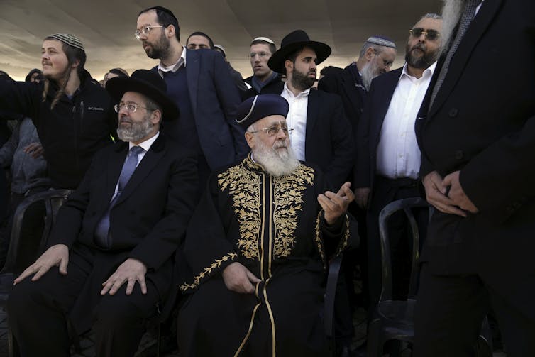 A crowd of men wearing head coverings, with one man seated in front wearing an ornate gold and black robe.