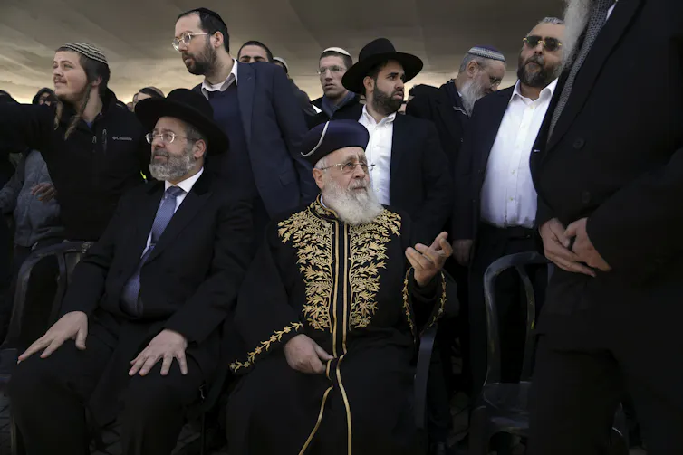 A crowd of men wearing head coverings, with one man seated in front wearing an ornate gold and black robe.