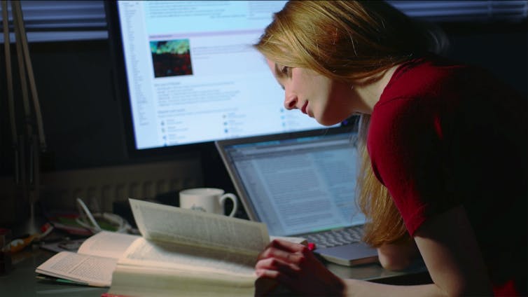 A woman peers into a book while seated at her desk, which has both a desktop and a laptop computer.