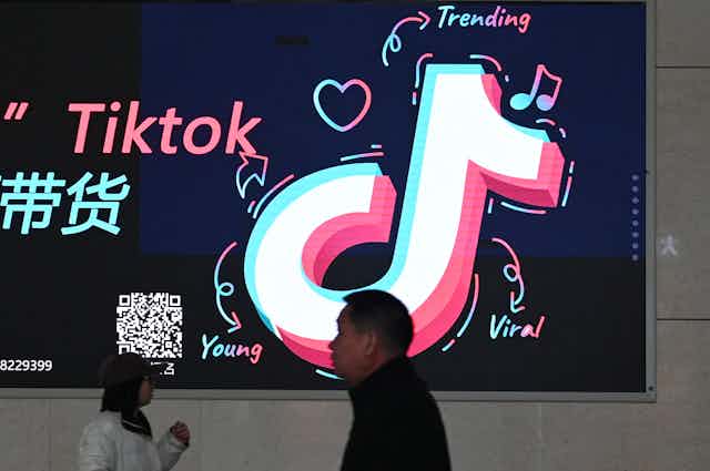 A man walks past a billboard with a neon symbol on it accompanied by Chinese writing.