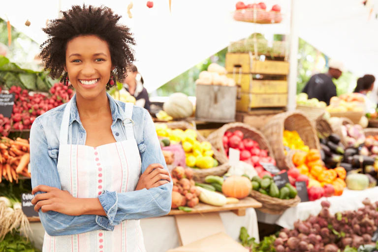 Black female stallholder with white apron at front left, market stall with fresh colourful fruit veg behind