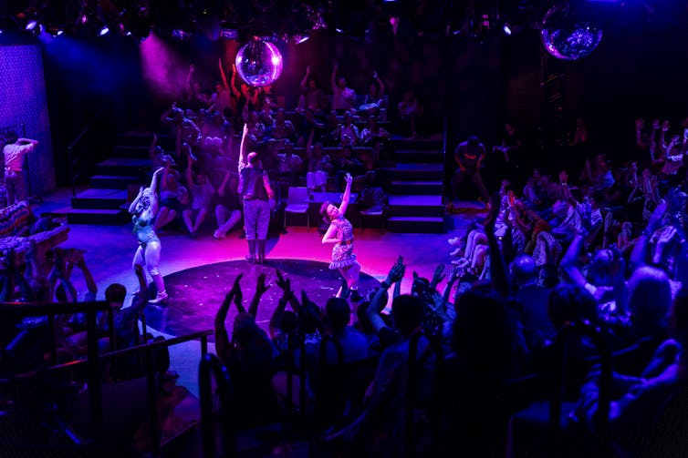The cast dance on a pink stage.