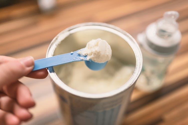 Woman holding blue plastic spoon of formula powder over open tin of formula, milk bottle in background