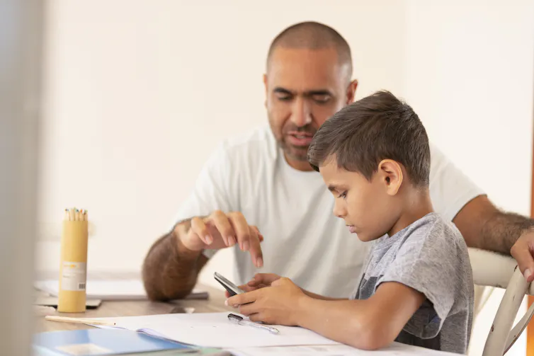 A young boy and an a man sit at a table looking at papers and a phone.