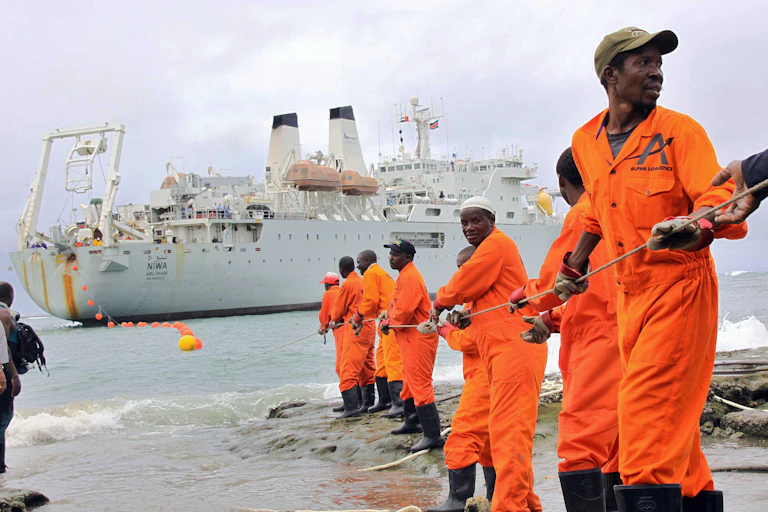 A group of men in orange overalls form a line to haul a cable to shore from a large ship