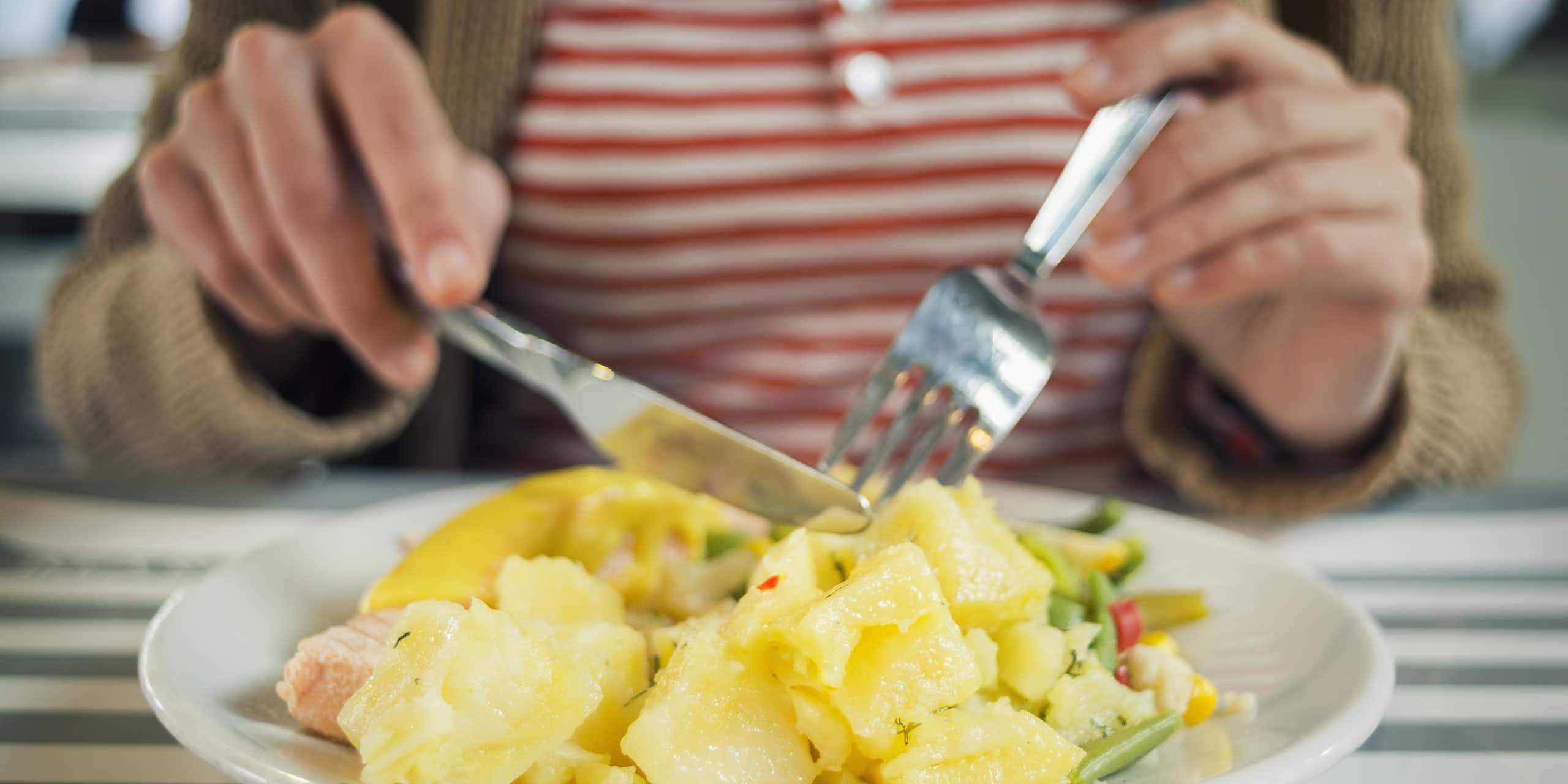 A woman uses a fork and knife to cut into a cooked potato.