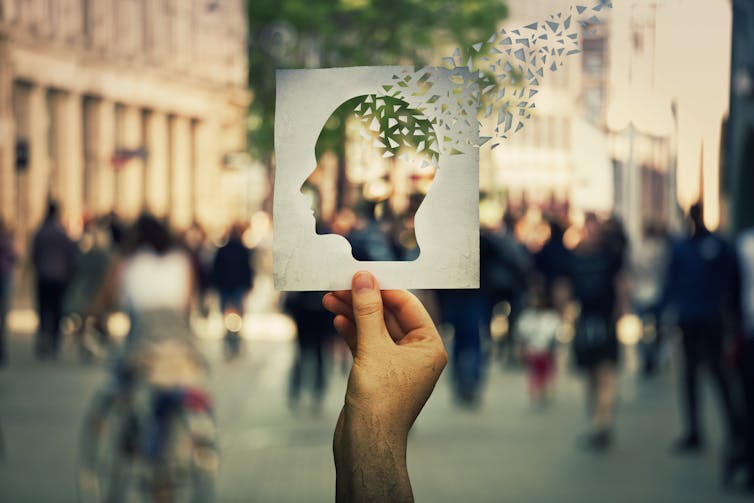 Hand holding a paper sheet with human head icon broken into pieces over a crowded street background.