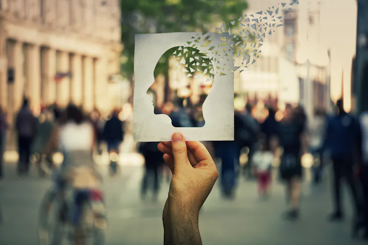 Hand holding a paper sheet with human head icon broken into pieces over a crowded street background.
