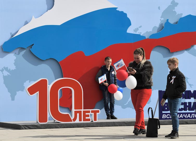 Boy and woman stand outside next to a large map of Crimea painted in Russian flag colours on a wall