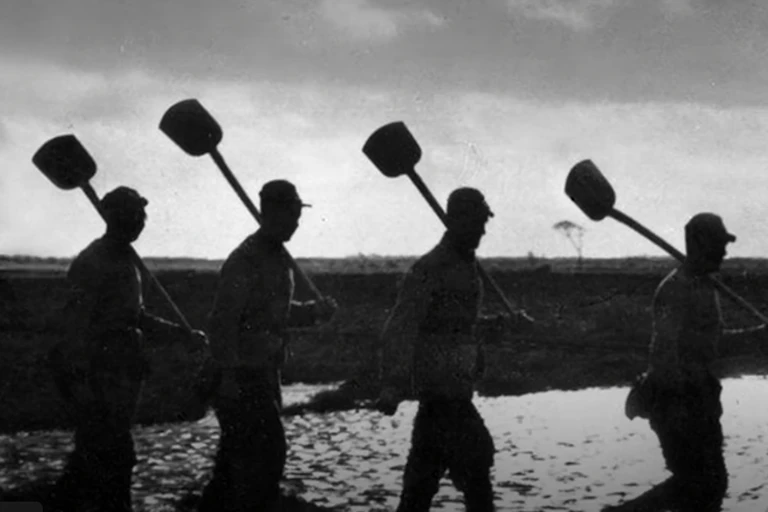 A group of men with shovels over their shoulders marching in a line silhouetted against the sky.