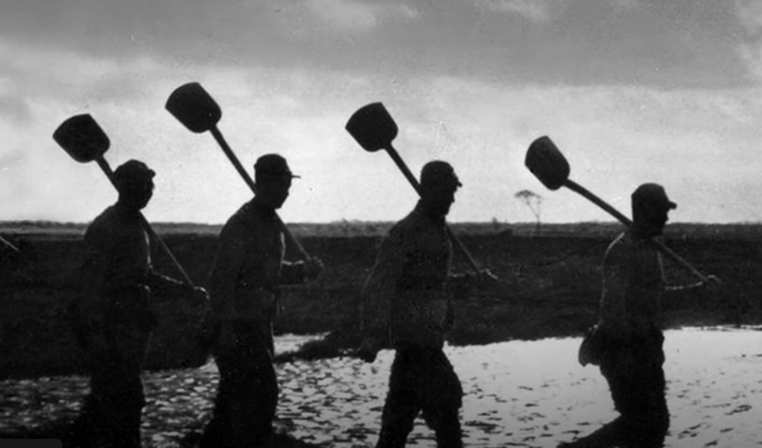 A group of men with shovels over their shoulders marching in a line silhouetted against the sky.
