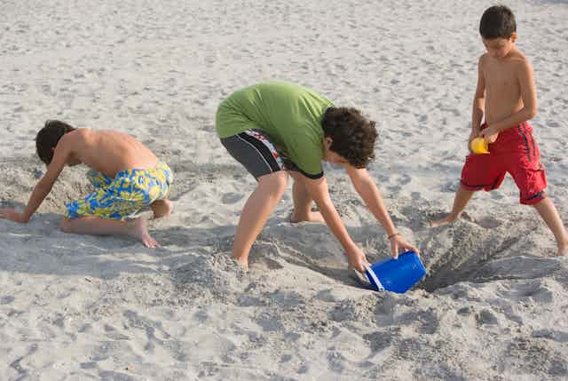Three young boys use plastic buckets and shovels to dig a hole in the sand.