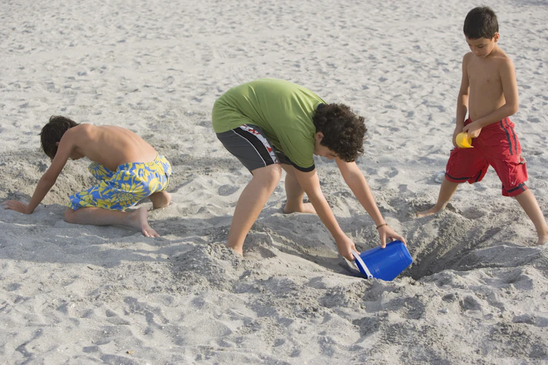 Three young boys use plastic buckets and shovels to dig a hole in the sand.