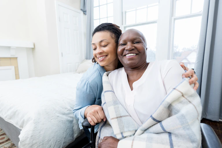 A smiling elderly woman in a wheelchair is hugged by her daughter.