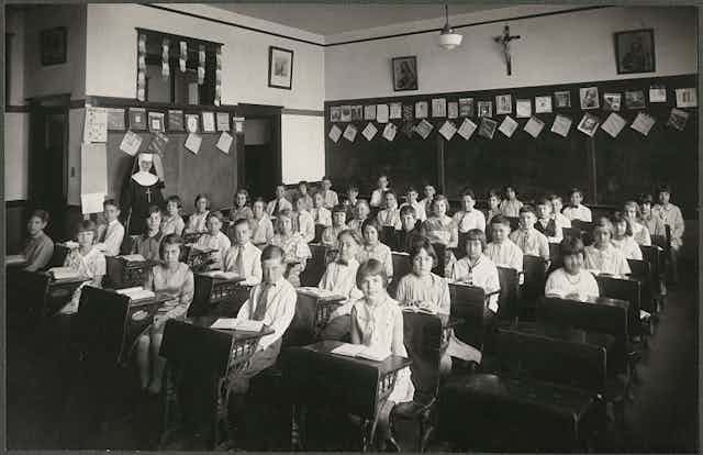 A black and white photograph shows a room with about 40 children seated at desks, and a woman in a nun's habit standing toward the back.