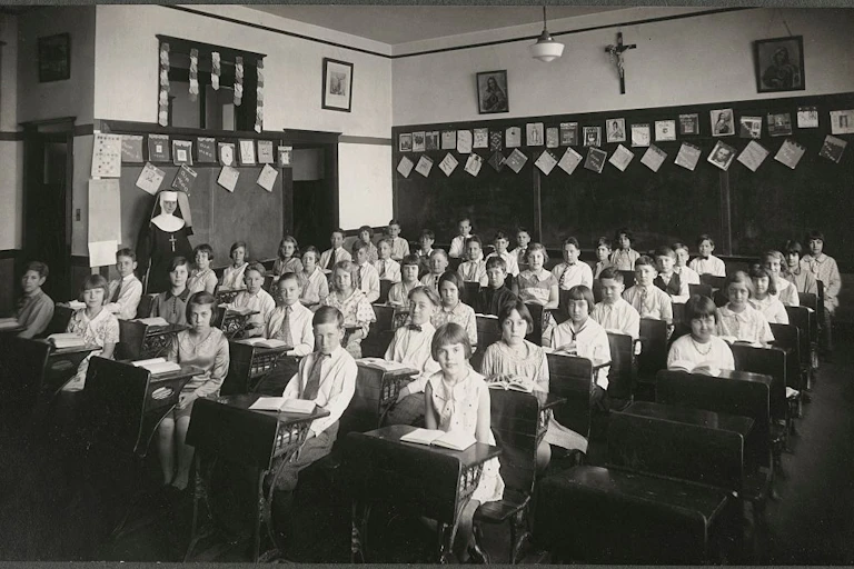 A black and white photograph shows a room with about 40 children seated at desks, and a woman in a nun's habit standing toward the back.