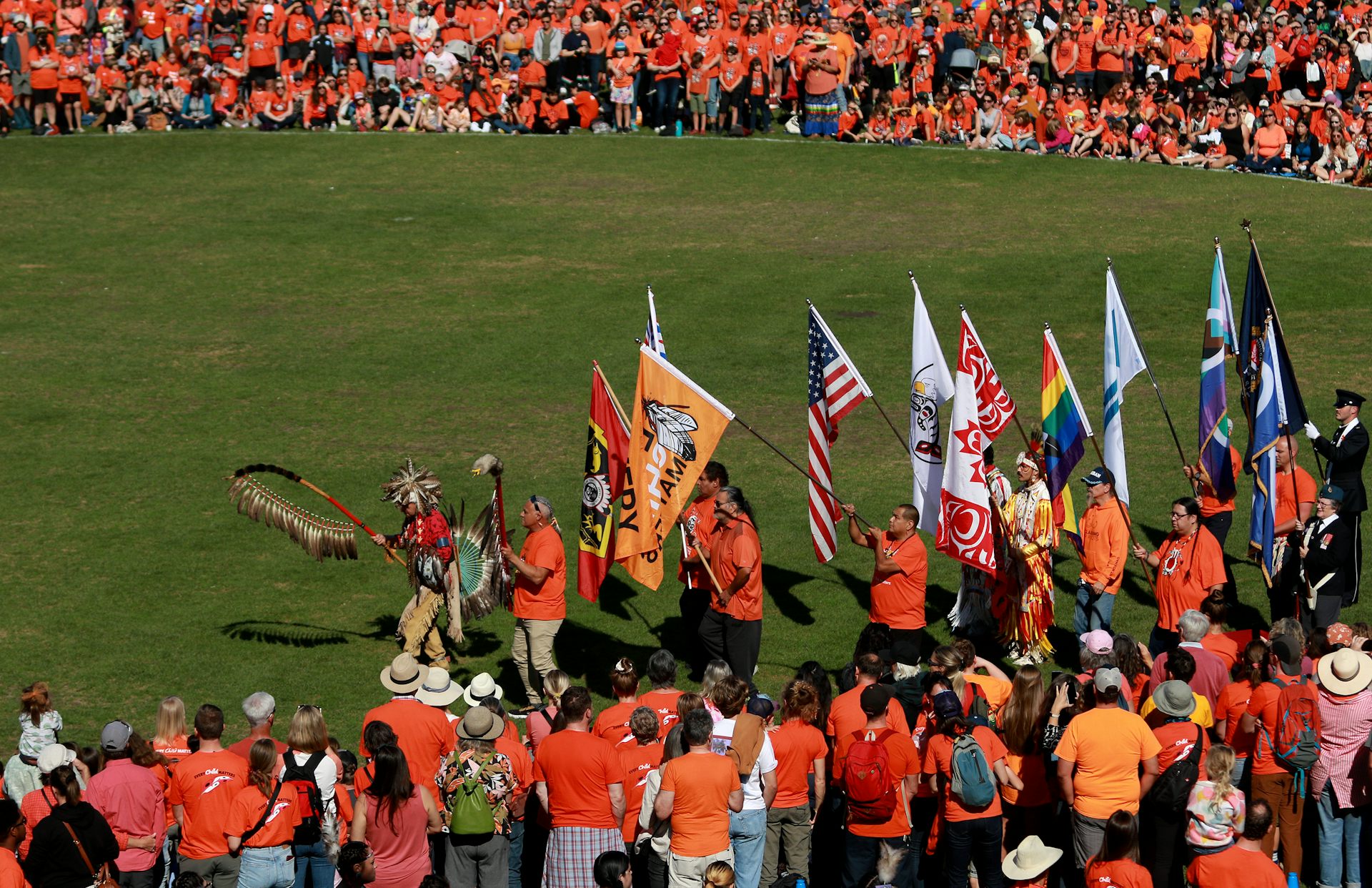 People seen in orange shirts a circle around with dancers in a line wearing powwow regalia.