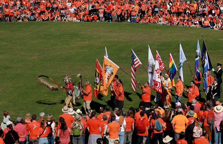 People seen in orange shirts a circle around with dancers in a line wearing powwow regalia.