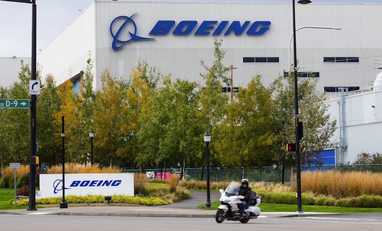 A motorcyclist drives past a building with the word Boeing on the side of it