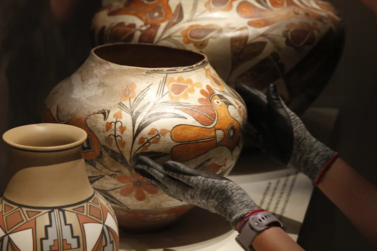 A pair of hands wearing gloves carry a vase painted with an orange bird and flowers
