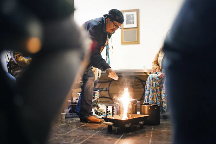 A man lights a small fire in a room with other people seated around.