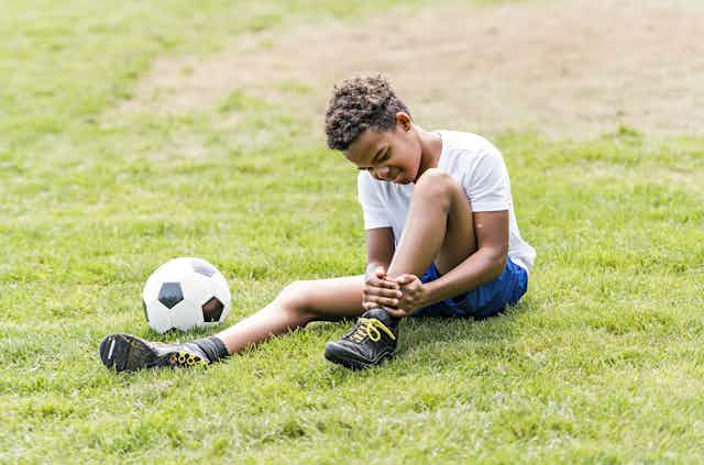 With a soccer ball nearby, a young athlete holds his ankle as he winces in pain.