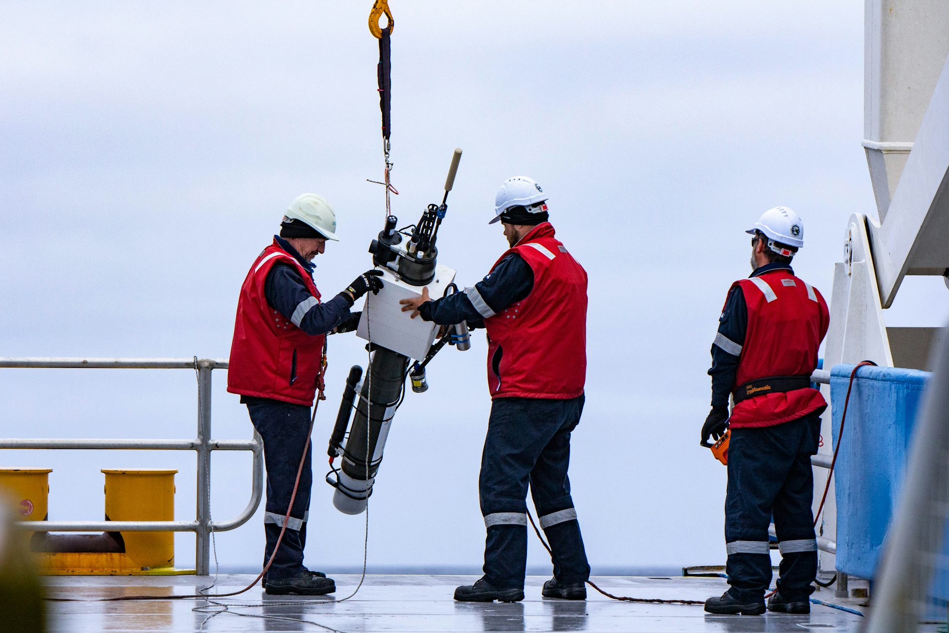 People in red safety vests handle a large machine while standing on the deck of a ship.