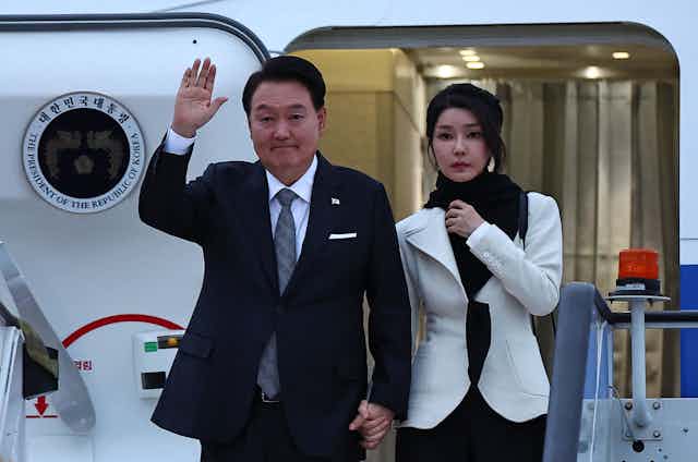 A man in a suit and a woman in white exit an airplane.