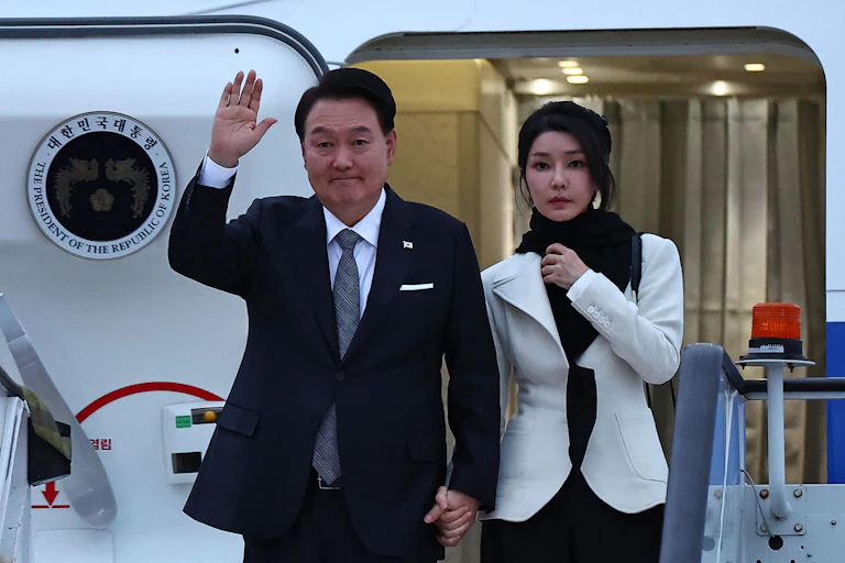 A man in a suit and a woman in white exit an airplane.