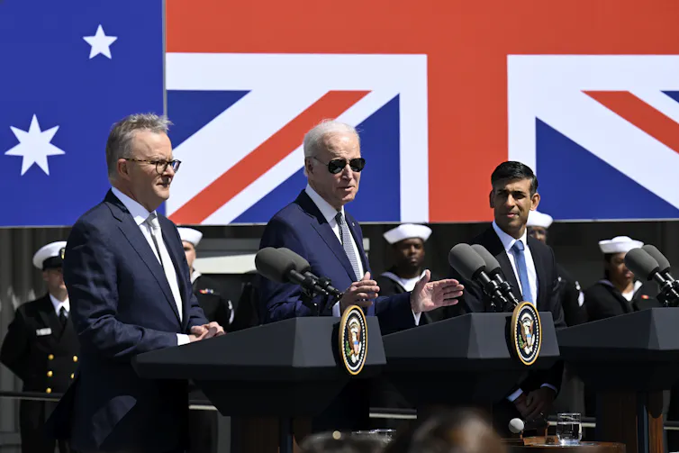 Three men stand behind lecterns in front of multiple national flags