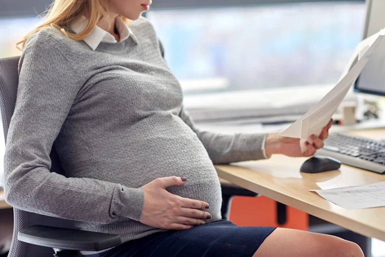 Woman sitting at desk reading from a document with one hand on her pregnant bellly