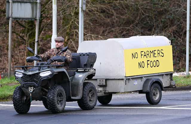 A man driving a quad bike, pulling a trailer with a sign on it that reads 'no farmers, no food'.