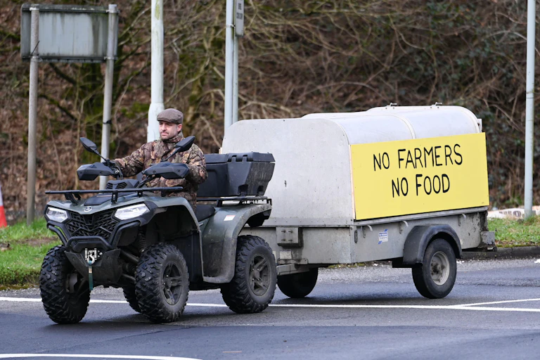 A man driving a quad bike, pulling a trailer with a sign on it that reads 'no farmers, no food'.