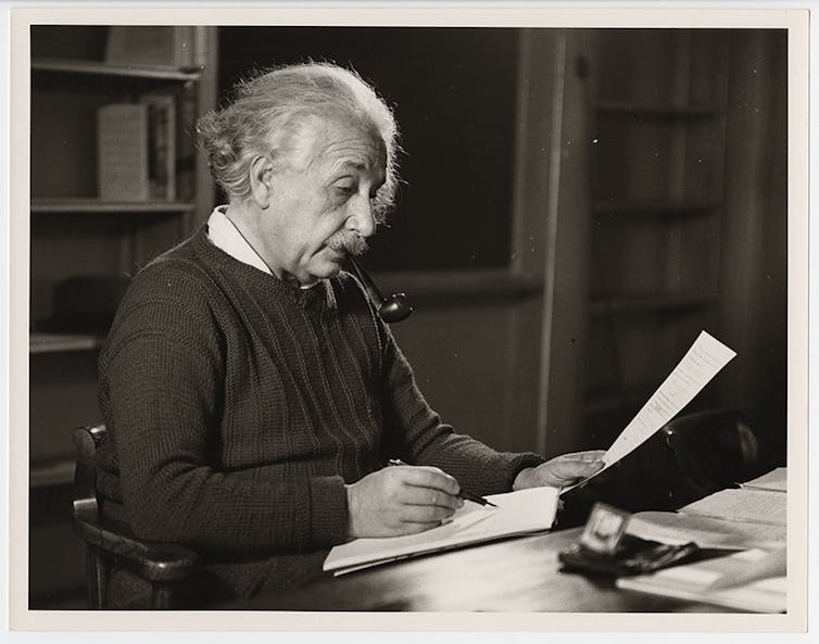 Albert Einstein sitting at his desk with pipe marking papers.