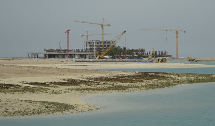 A distant construction site with cranes with sand and shallow water in the foreground.