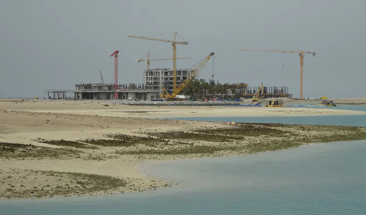 A distant construction site with cranes with sand and shallow water in the foreground.