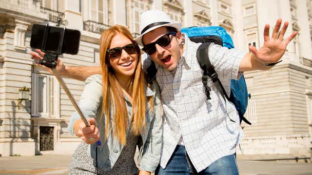 A young tourist couple in sunglasses taking a selfie with a selfie stick, in front of a grand building