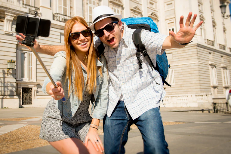 A young tourist couple in sunglasses taking a selfie with a selfie stick, in front of a grand building