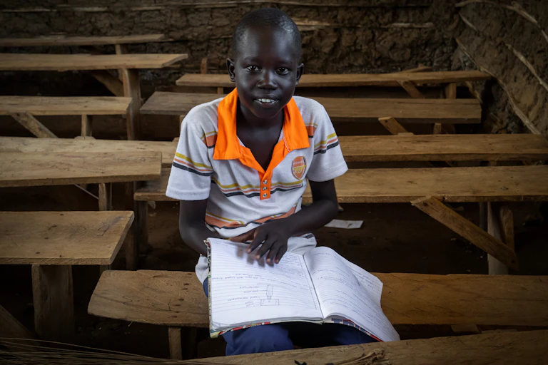 Young black student boy in white/orange T shirt sat with open white book on wooden bench, empty wooden tables and chairs around him, dark background