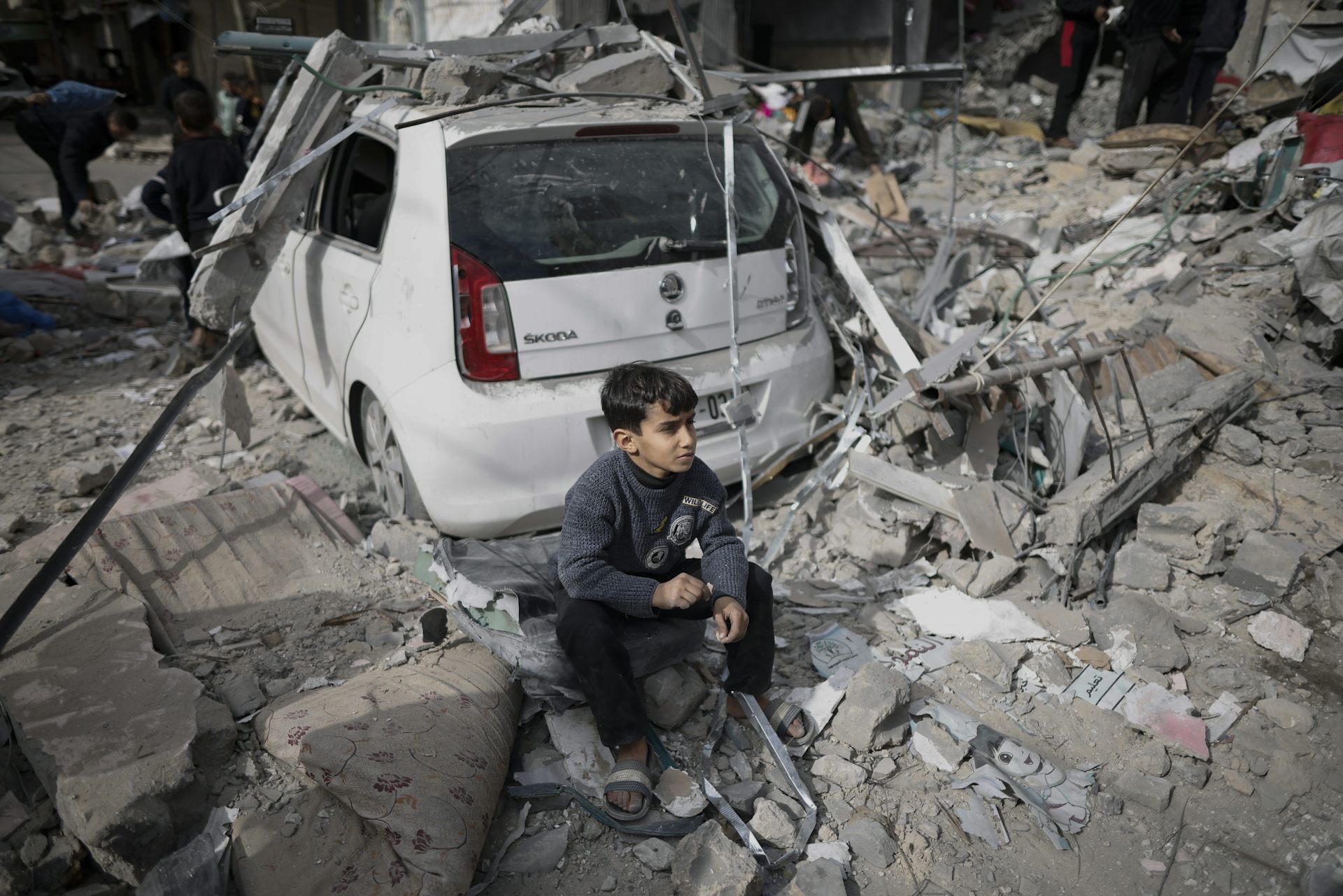 A boy looking despondent sits near a car in front of rubble where his home used to be.