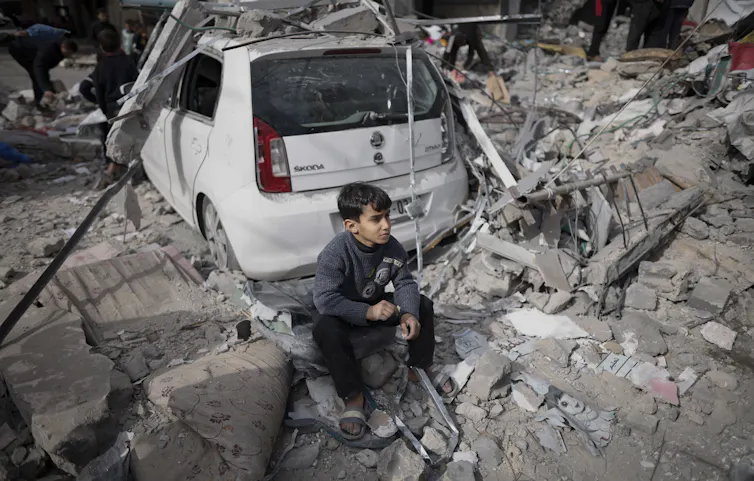 A boy looking despondent sits near a car in front of rubble where his home used to be.