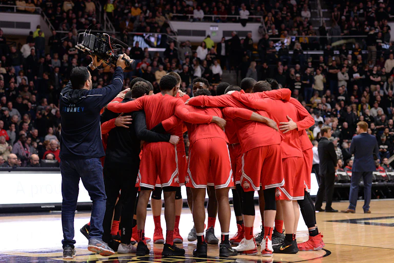 Several college basketball players clad in scarlet are seen on a court; behind them, a cameraman wearing dark, unobtrusive colors angles for a shot.