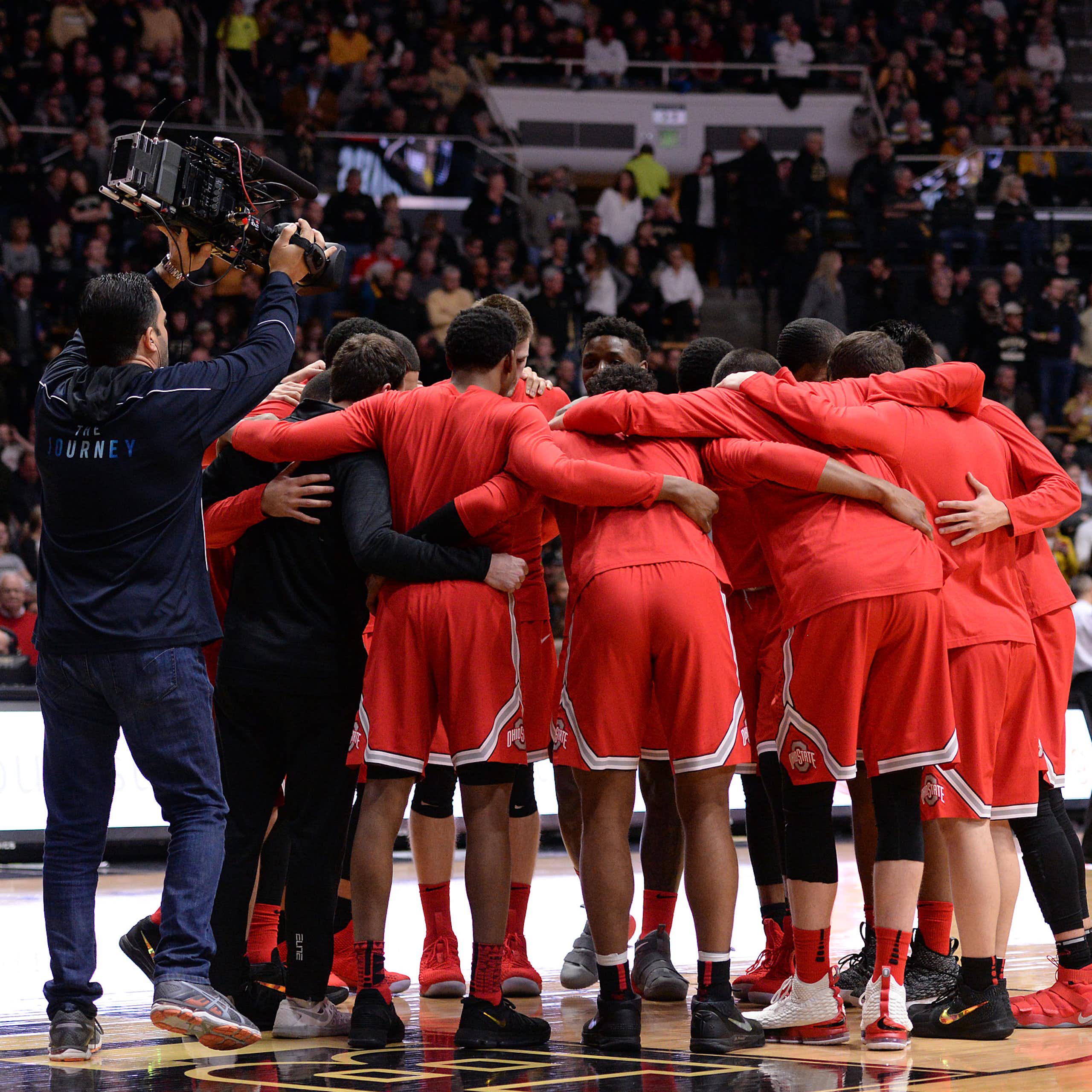 Several college basketball players clad in scarlet are seen on a court; behind them, a cameraman wearing dark, unobtrusive colors angles for a shot.